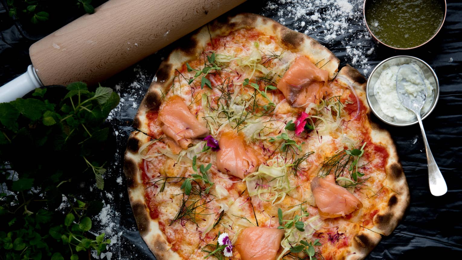 A wood-fired pizza topped with smoked salmon, fennel and dill, bowls of green and white sauces on the side. 