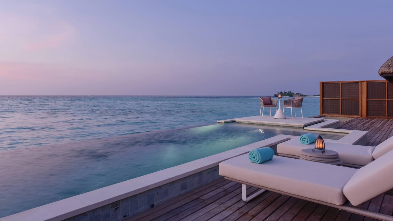 A lounging area next to the water with lounge chairs and a clear sunsetting sky.