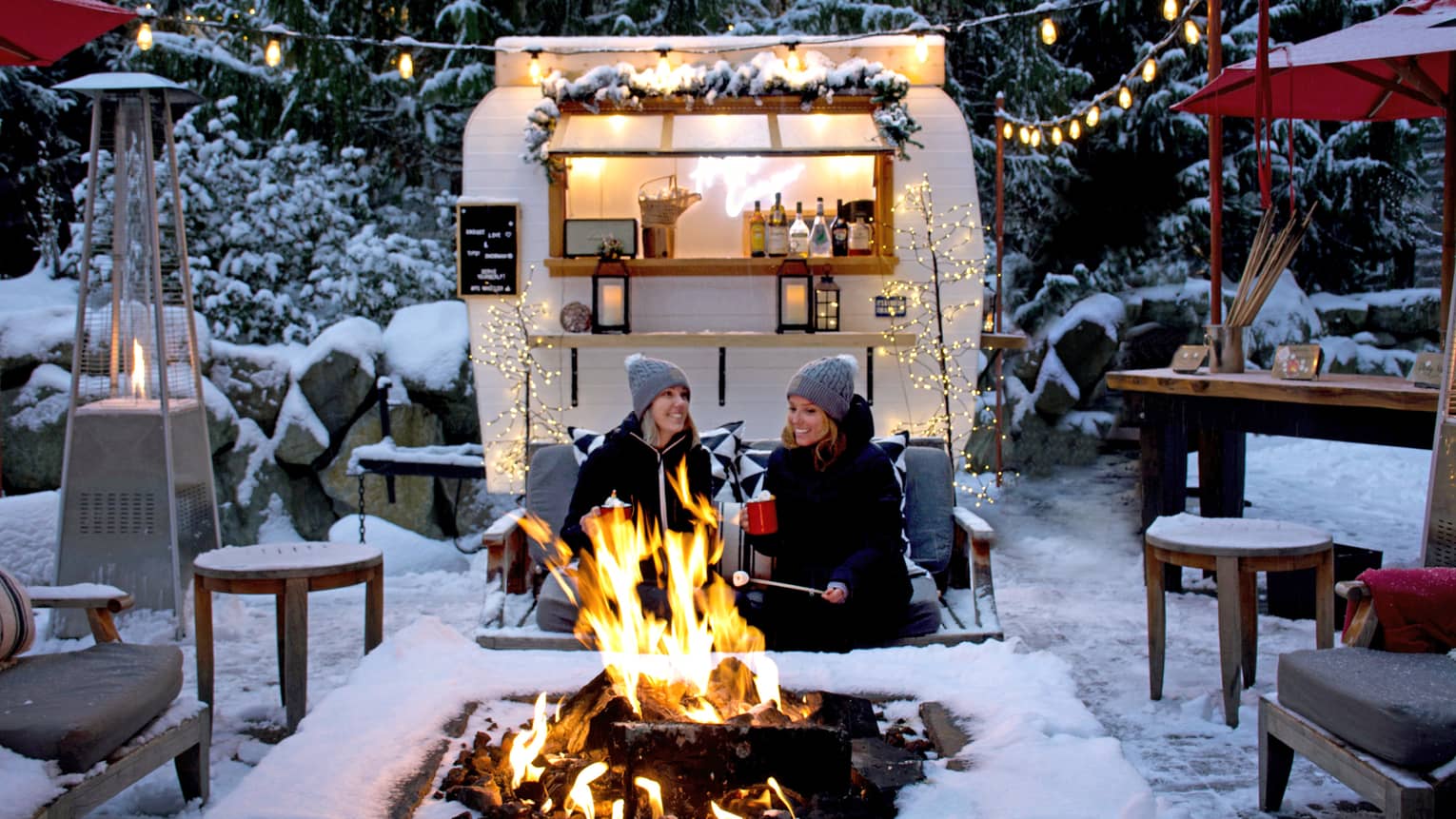 Two women roasting marshmallows at an outdoor fire pit with camper in background