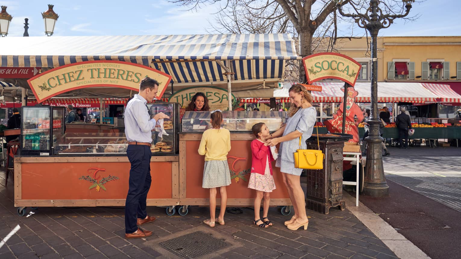 Man in blue shirt, black pants with two girls and woman in dresses at market farm stand)