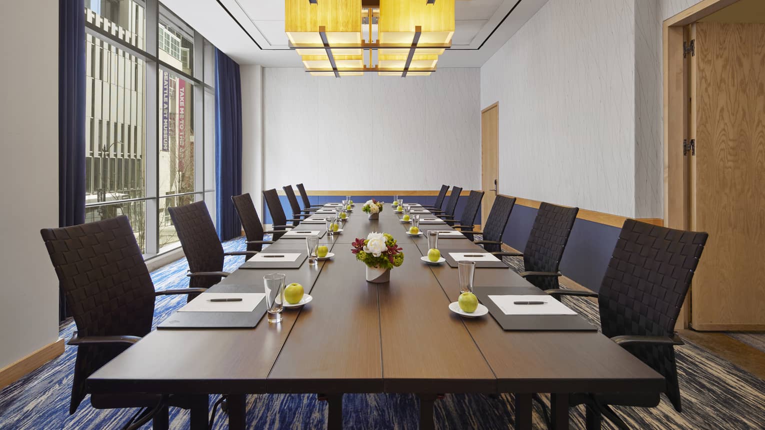 A room with a long table with black chairs on two sides and a large window on one wall.