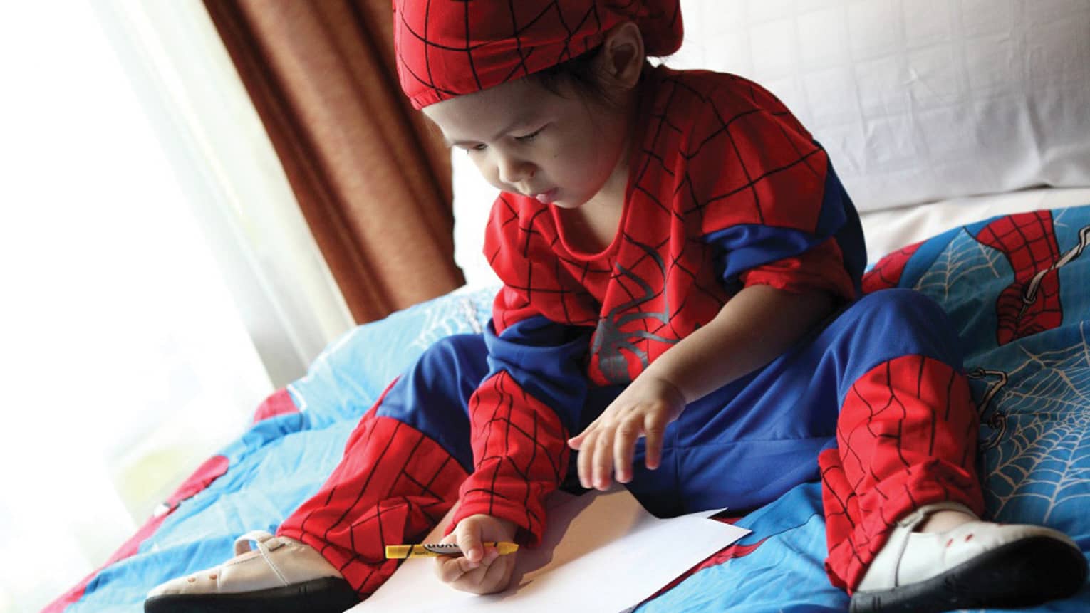 Young child wearing Spiderman costume sits on bed, colours in book