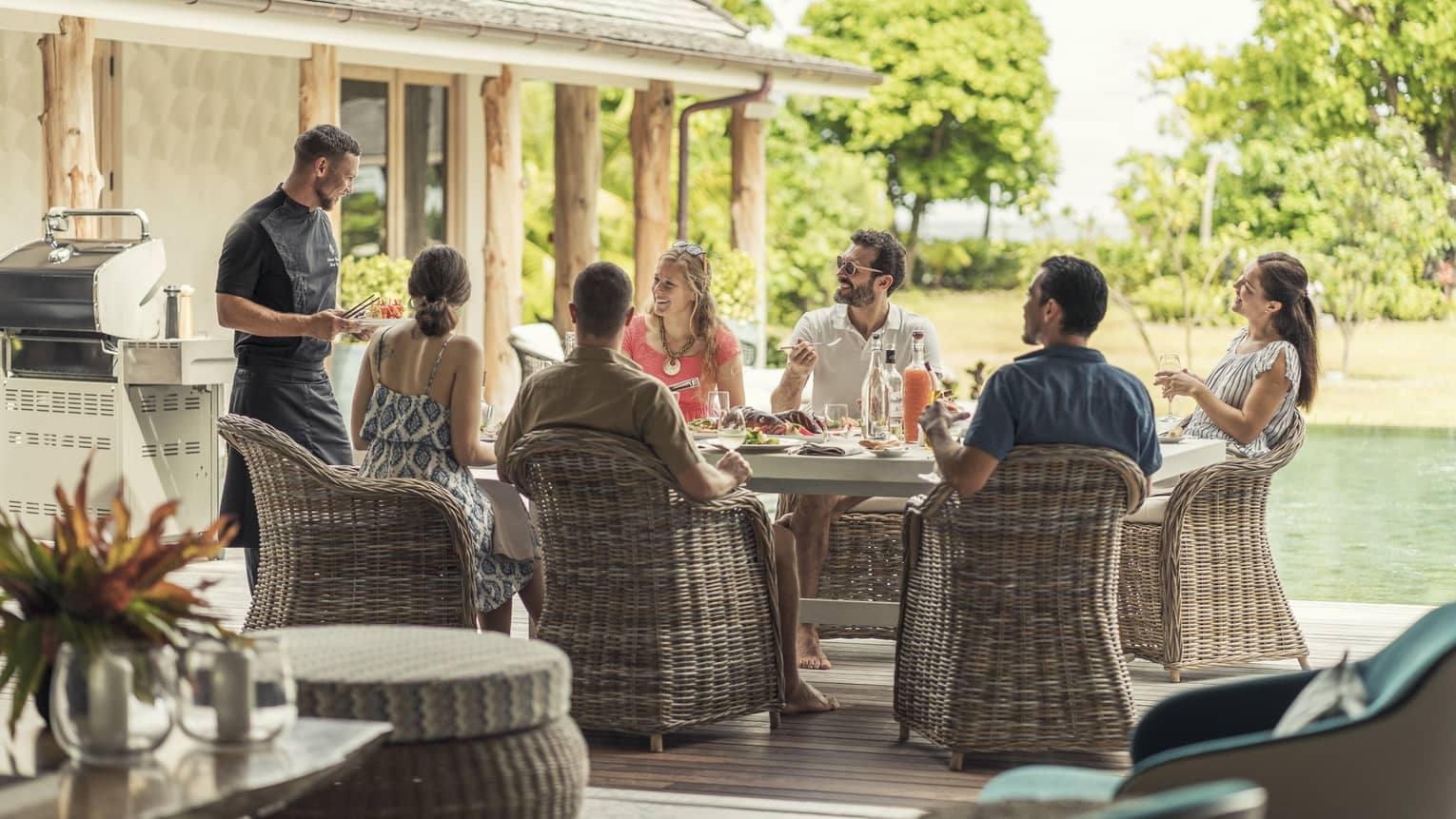 Group of men and women on wicker chairs by pool eating and talking to chef