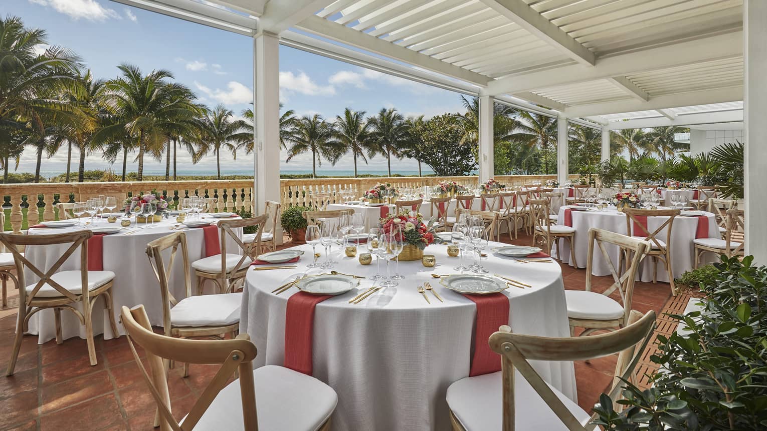 Elegantly set round dining tables and chairs on white veranda with view of palm trees and ocean