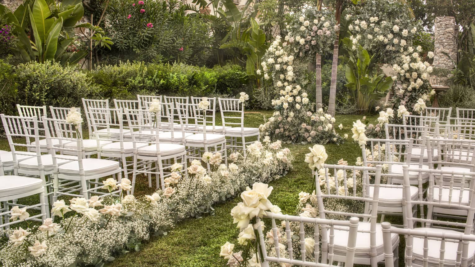 Rows of white dining chairs lined up along floral aisle leading to flower arch amid garden wedding setup