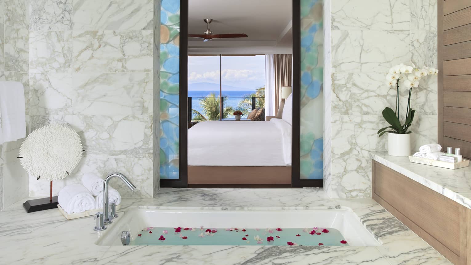 White marble bathroom with water- and rose petal–filled soaking tub and view through bedroom