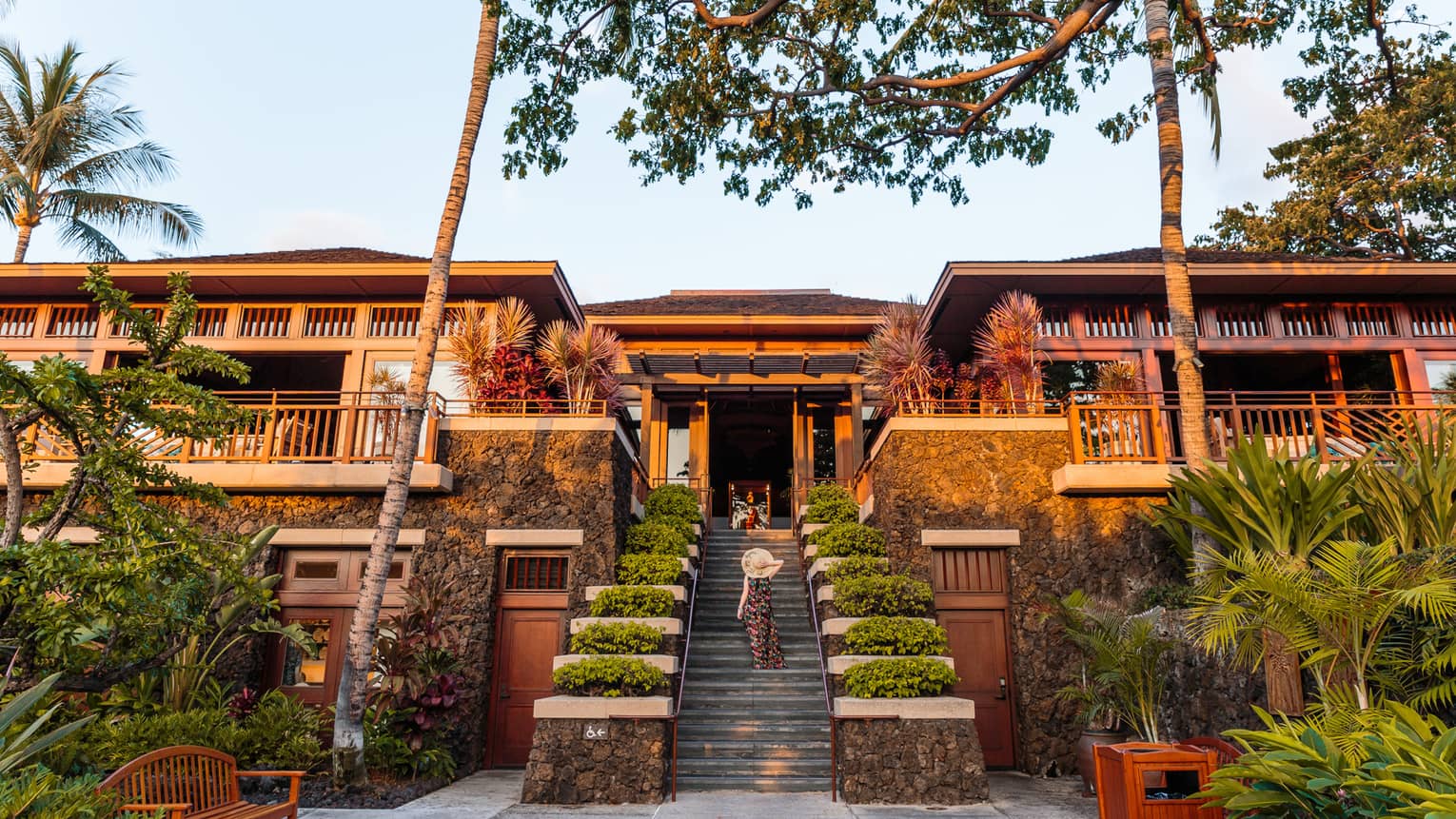 Woman in sunhat walking up resort stairs with shrubs, two-storey building with balconies