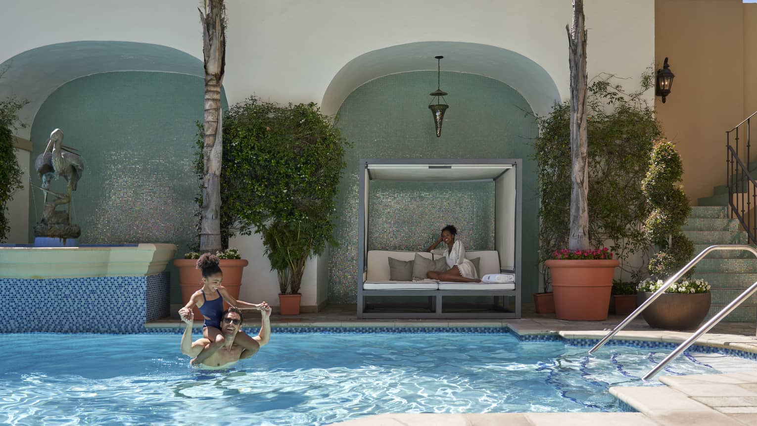 A father and daughter in the pool while a mother sits on a lounge chair.