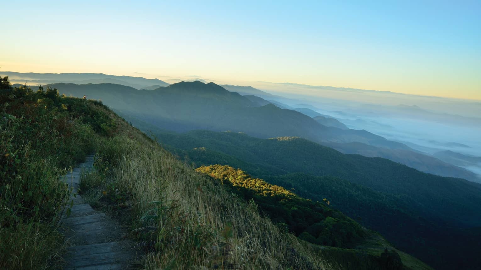 View from an elevated grass-lined path of low, forested mountains receding into the misty distance under blue and orange sky.