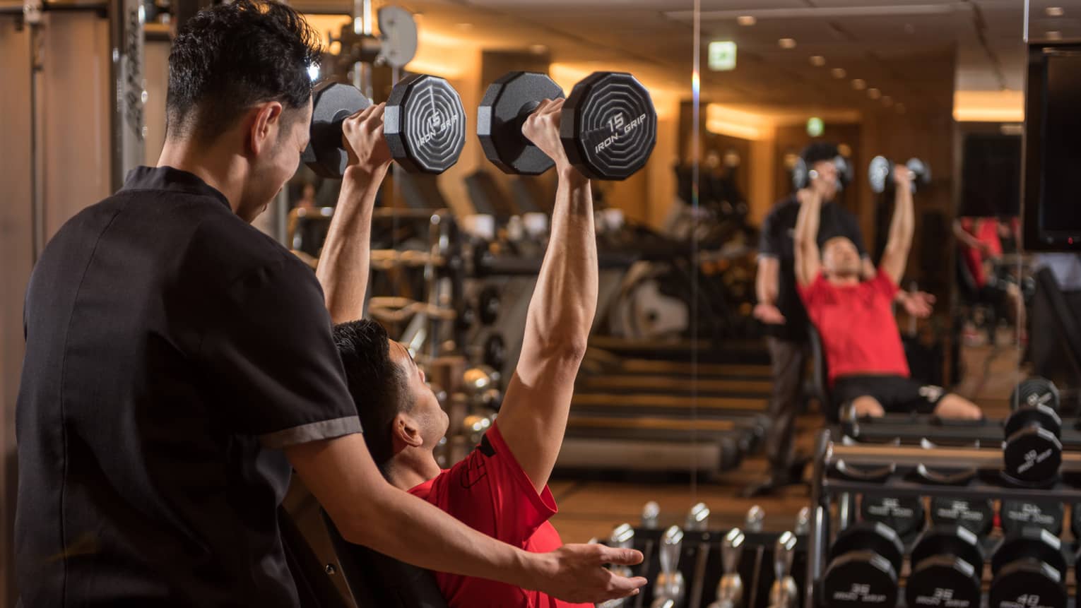 Man in chair in front of mirror lifts two 15 lb. weights above his head while personal trainer assists