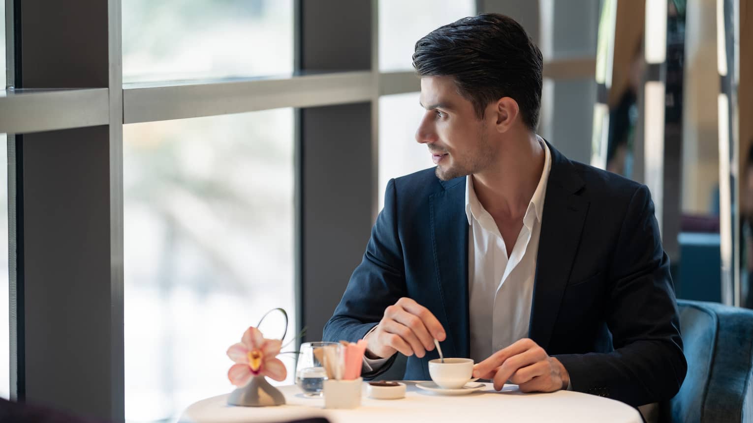 Man wearing black suit and white collared shirt with no tie sits at a table stirring a cup of esspresso while looking out the window
