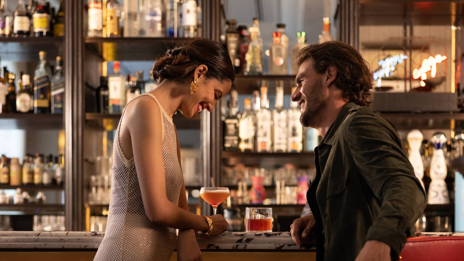 Elegantly dressed couple enjoy unique cocktails at a luxurious, dimly lit bar, with bottles of liquor on mirrored shelves. 