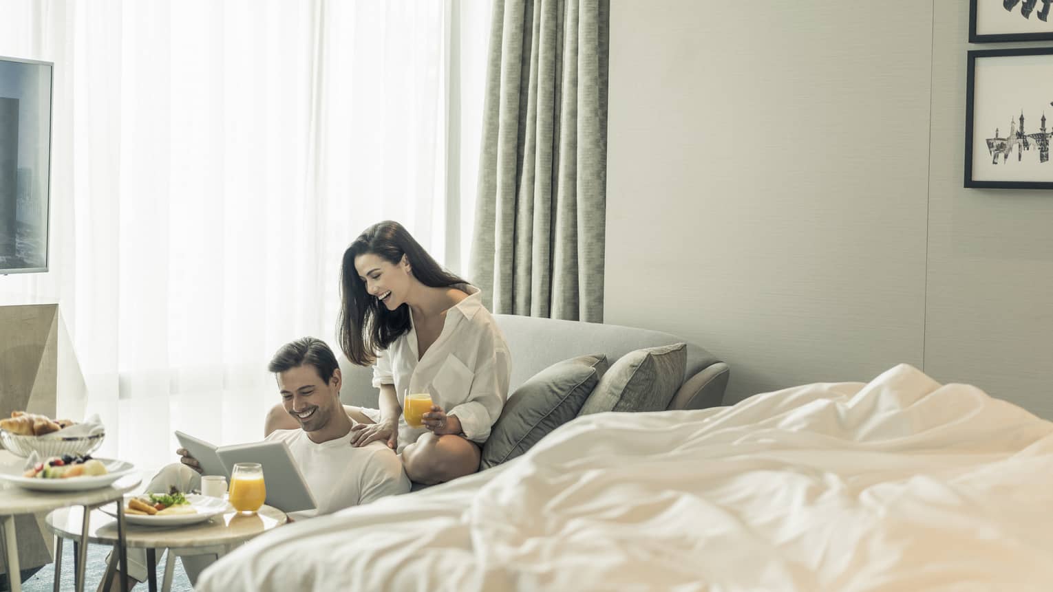 Couple wearing pyjamas enjoy in-room dining breakfast at window beside unmade bed 