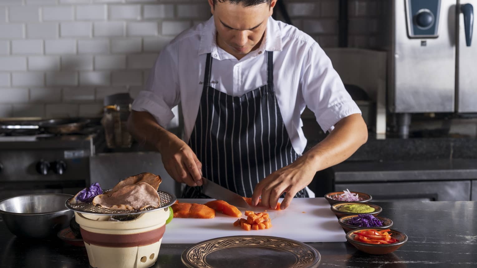 Chef wearing white button-down shirt and a navy-and-white striped apron slices salmon on a white cutting board in an industrial kitchen