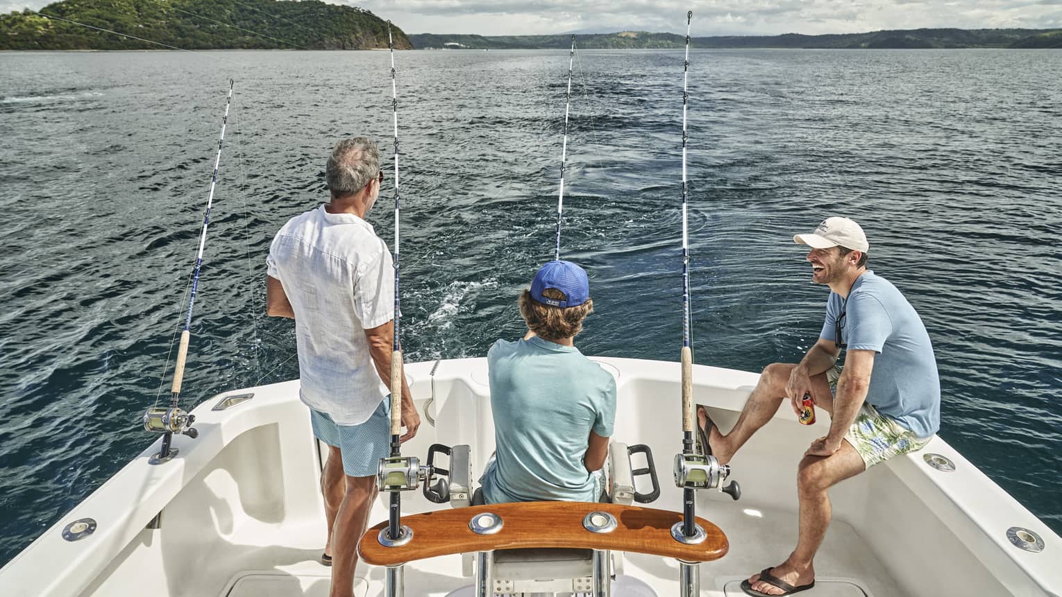 Three men of varying ages sit looking out at the water from the back of a boat outfitted with fishing rods