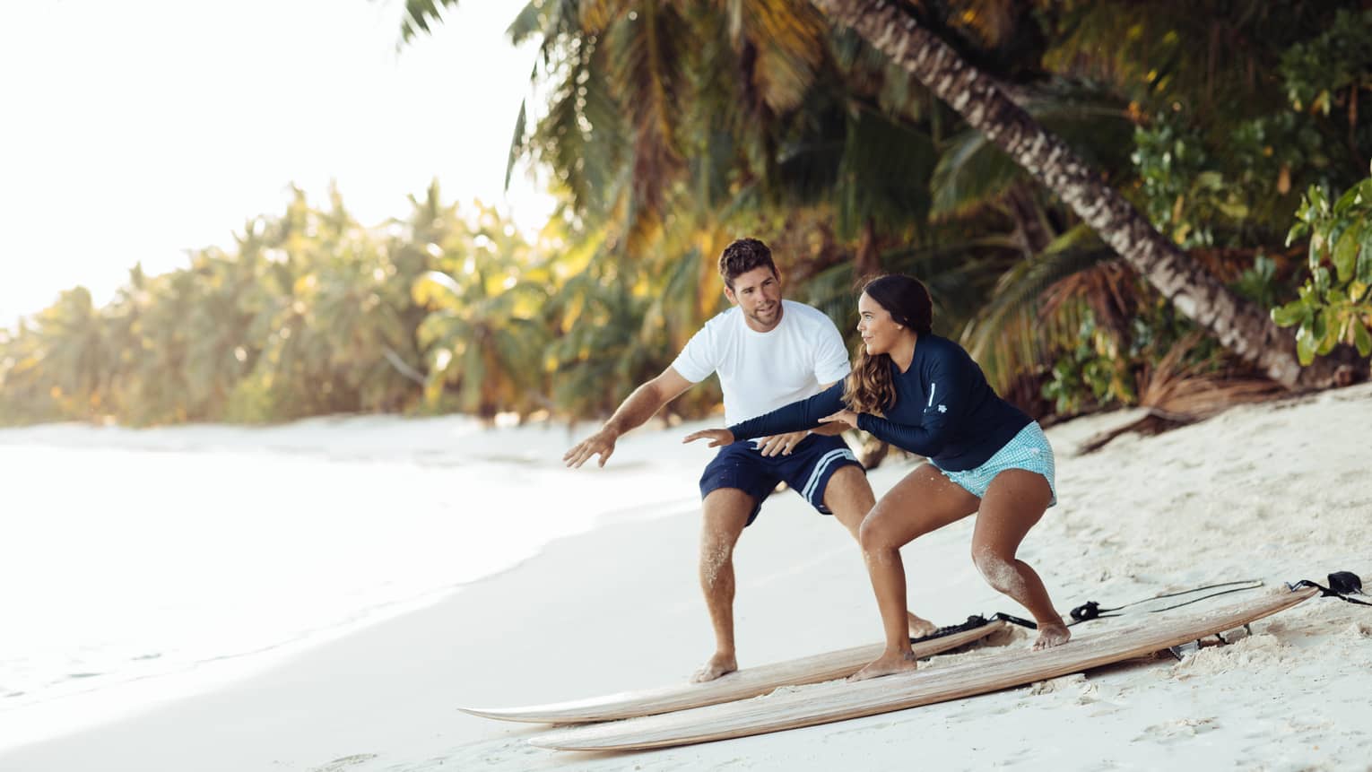 Man shows woman a surfing pose as they balance on surfboards on white sand beach under palm tree
