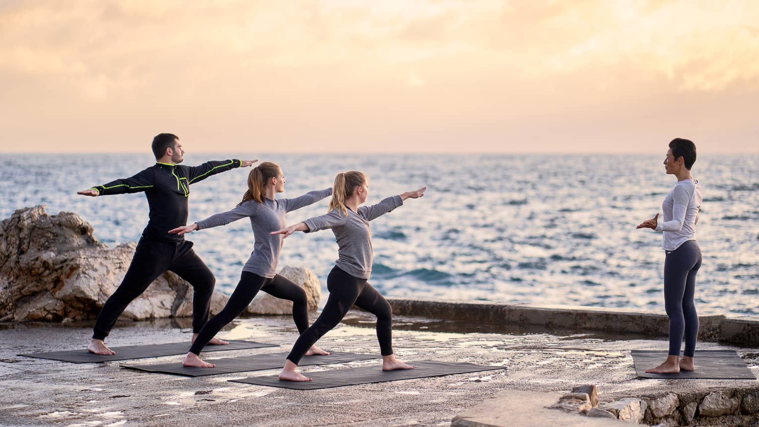 Three people in warrior yoga pose and coach standing at sea's edge at sunrise