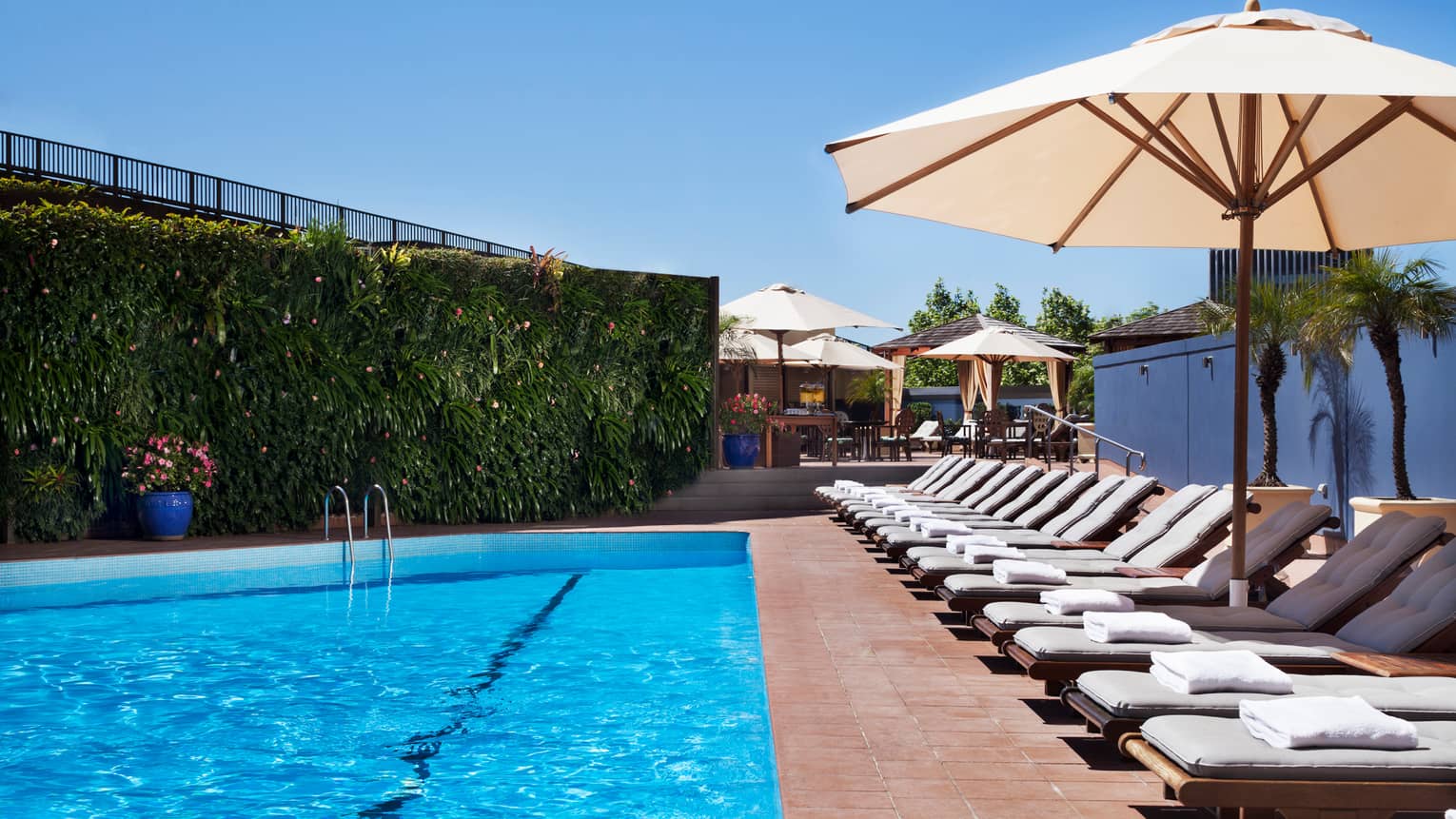 White lounge chairs lined up on deck under patio umbrella by blue outdoor swimming pool