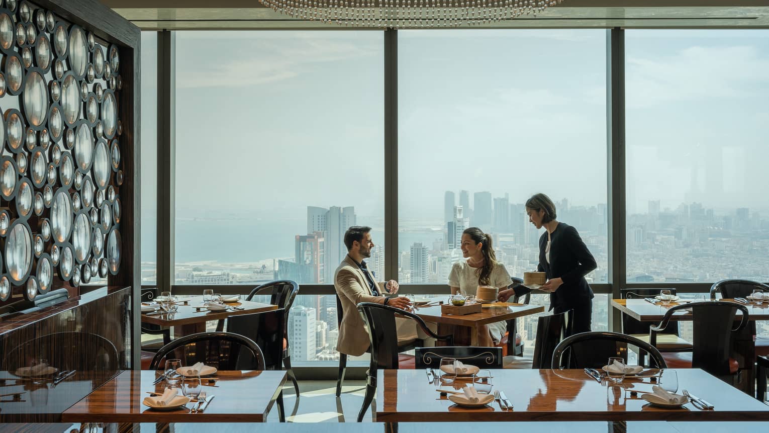 Several people meet in an Art Deco dining room overlooking the Bahrain cityscape. 