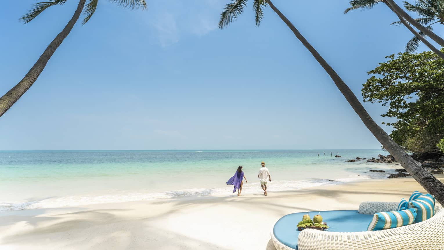 Couple walks along the shoreline framed by palm trees, round turquoise lounge chair in the foreground