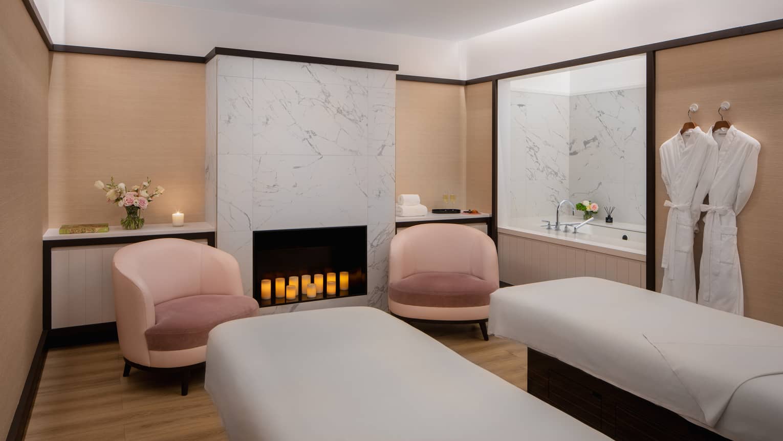 Couple's spa suite with a white-tiled fireplace filled with lit candles, an inset bathtub, two pink arm chairs, two side-by-side treatment tables and a pair of white robes