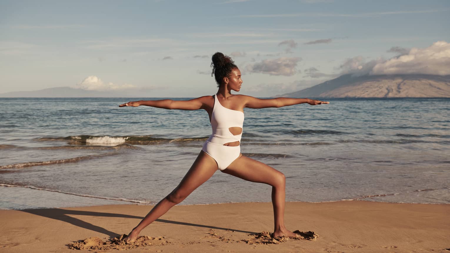 Woman wears white swimsuit and does warrior yoga pose on Maui beach