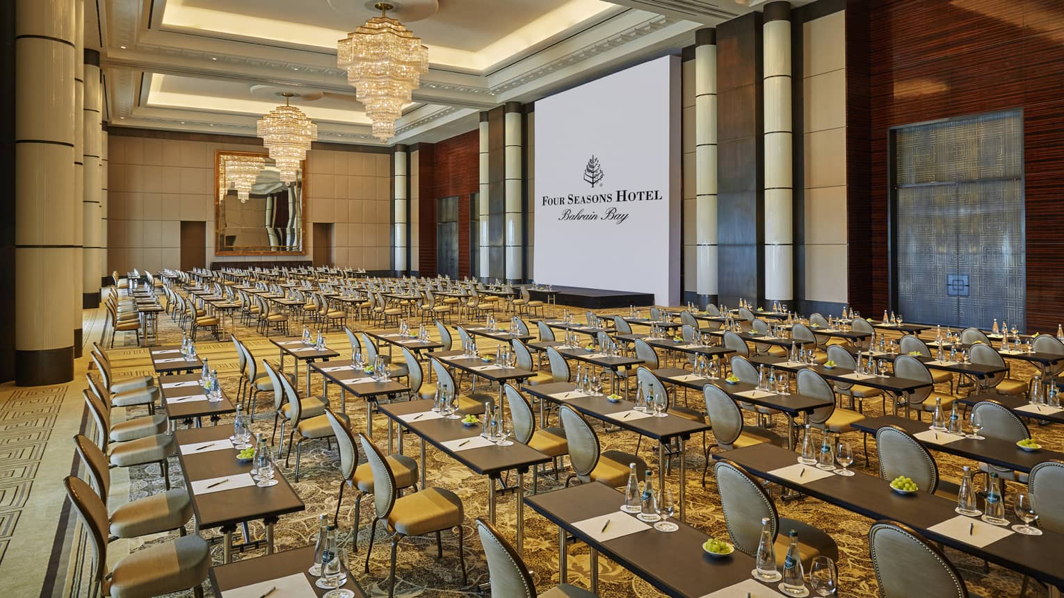 Conference in ballroom, rows of meeting tables and chairs facing large screen with Four Seasons Hotel logo