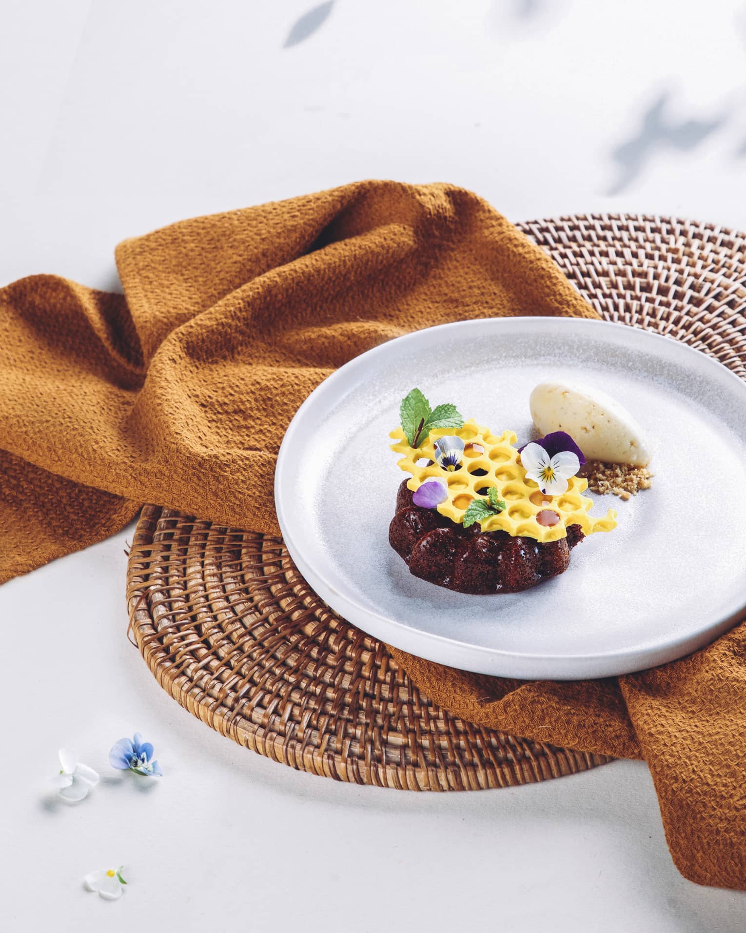 A rich chocolate mini-cake topped with honeycomb-shaped white chocolate garnished with mint leaves and dainty flower petals