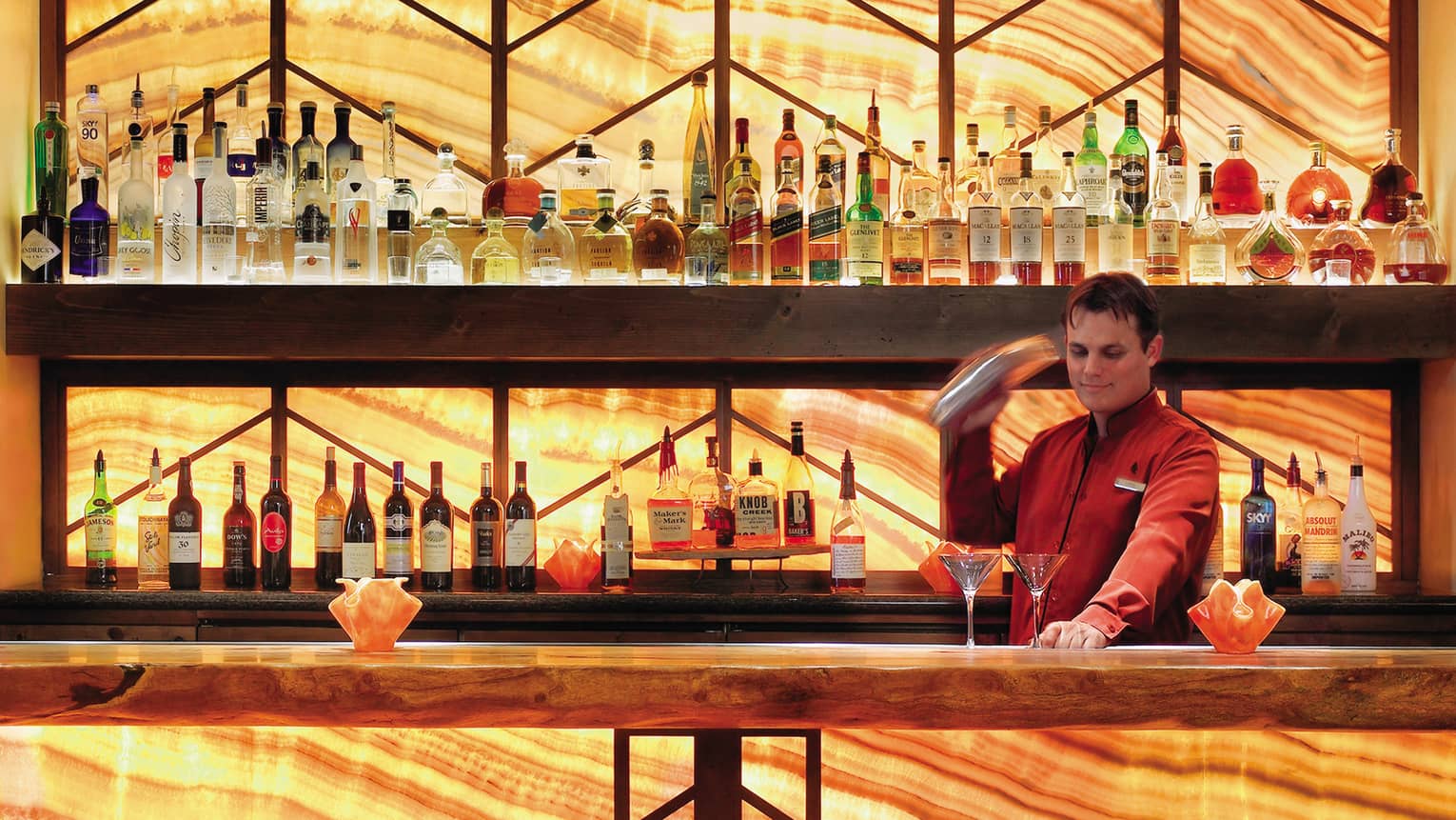 Bartender in red jacket with cocktail shaker in front of two martini glasses on bar with illuminated wood grain