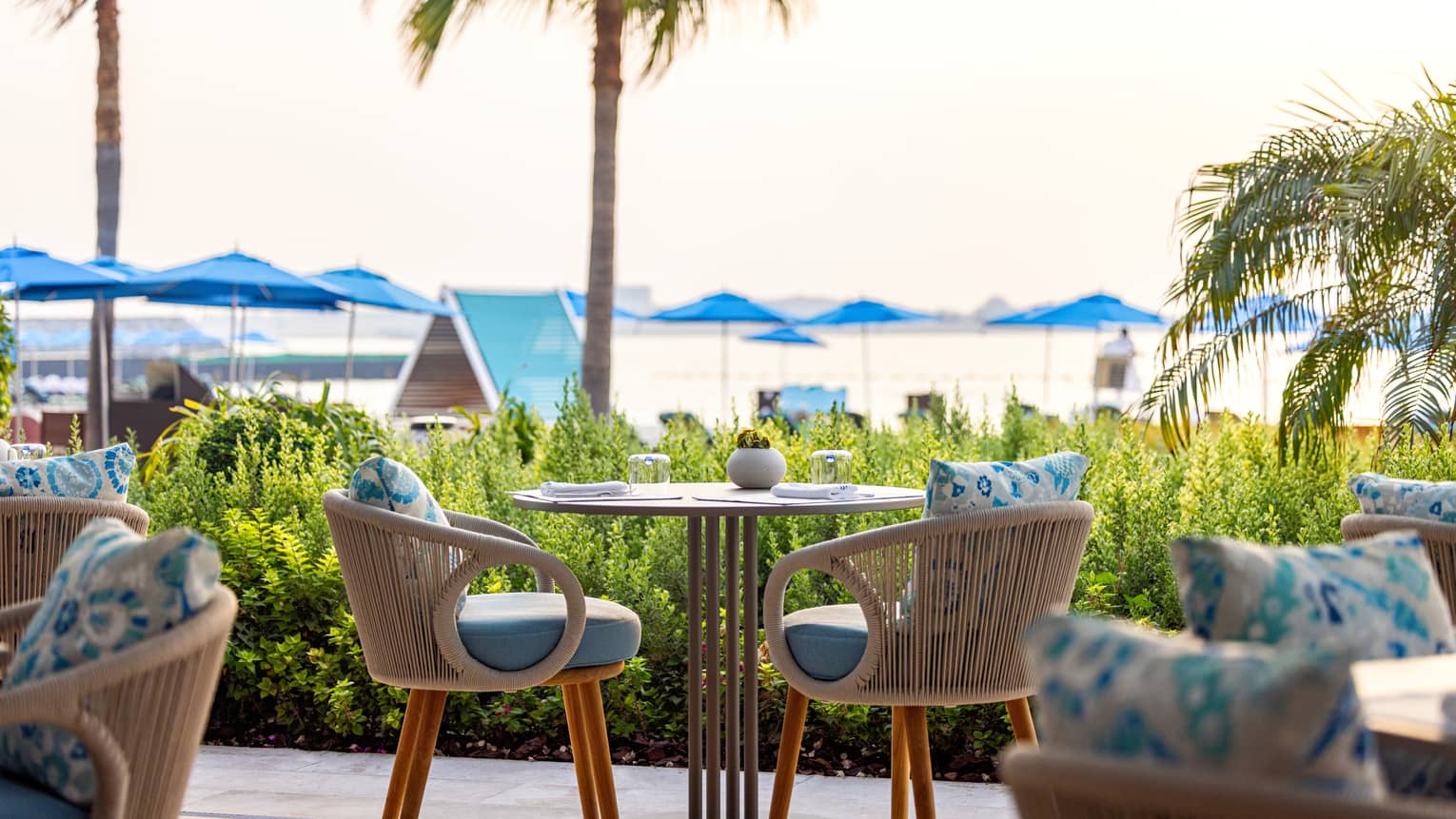 Small table for two set on an open-air terrace overlooking a lawn with palm trees and beach umbrellas in the distance