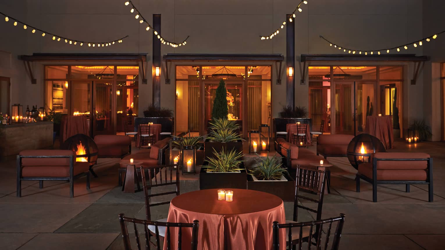 Terra courtyard at night, lounge furniture surrounding fire pits, candles, strings of lights 