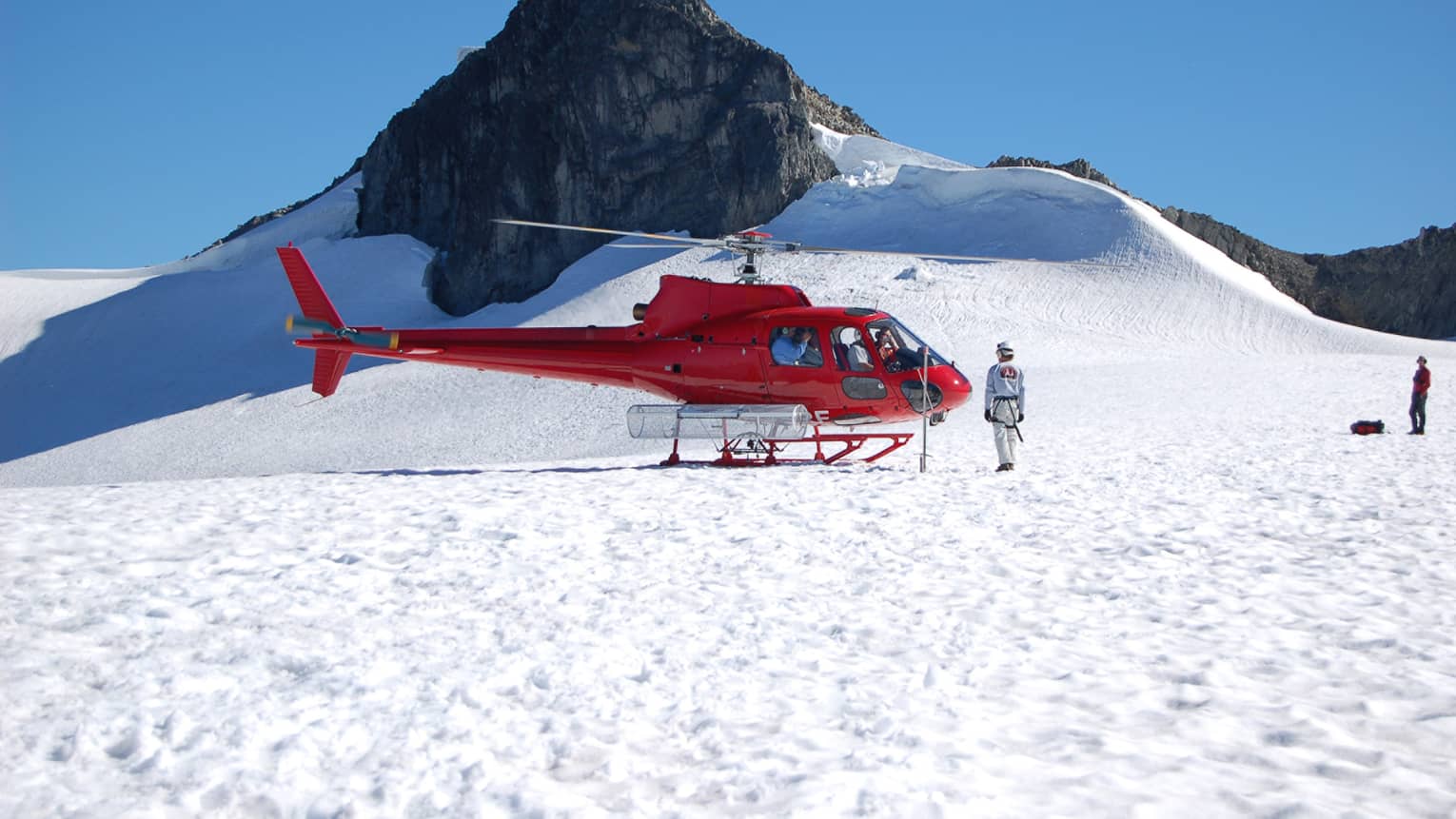 Person stands in front of a red helicopter landed on top of a glacier, surrounded by blue sky