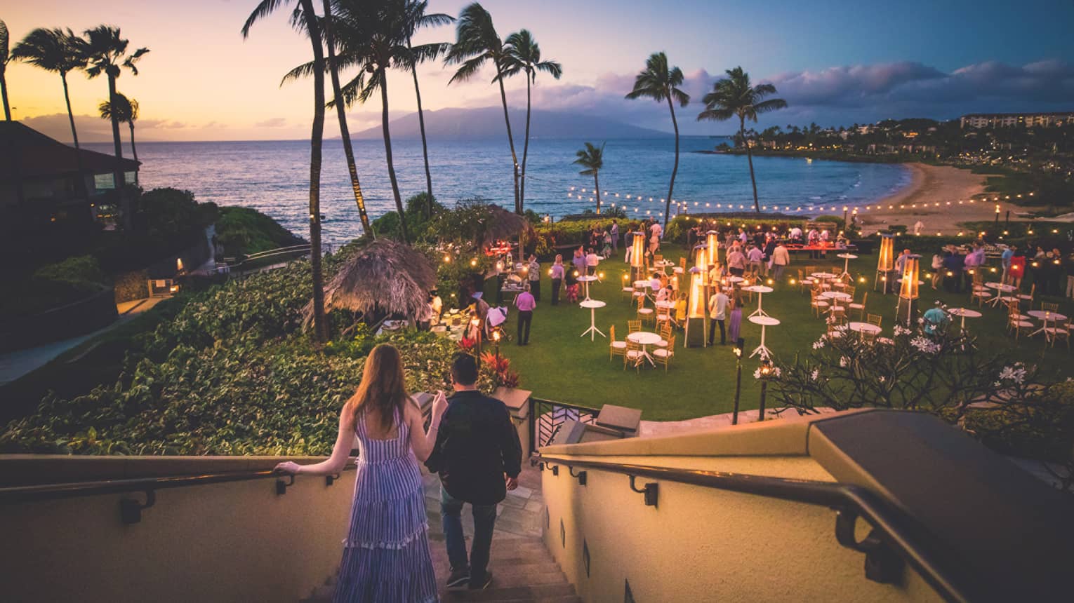 Couple walk down stairs towards grass with tables, lantern, surrounded by palm trees and ocean at dusk