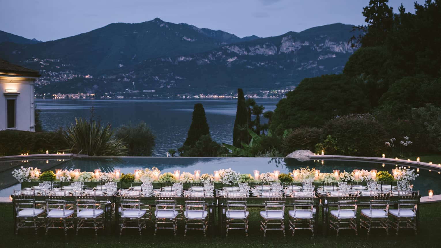 Long outdoor dining table with flowers, glowing candles by Lake Como, mountains at dusk