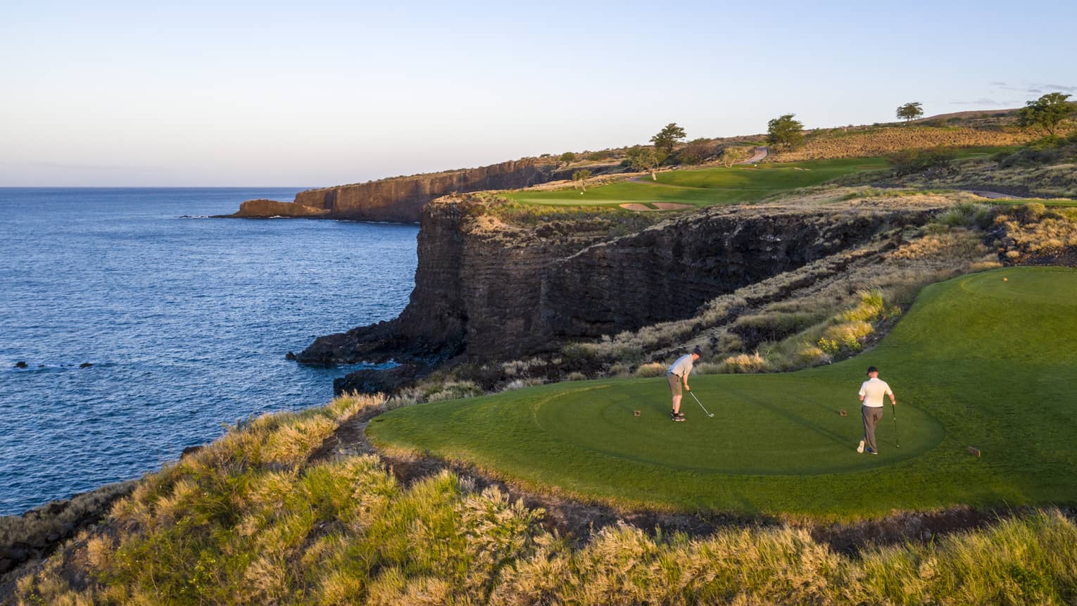 Two golfers at the 12th-hole tee at Manele Golf Course at Four Seasons Resort Lanai, cliffs and ocean in background