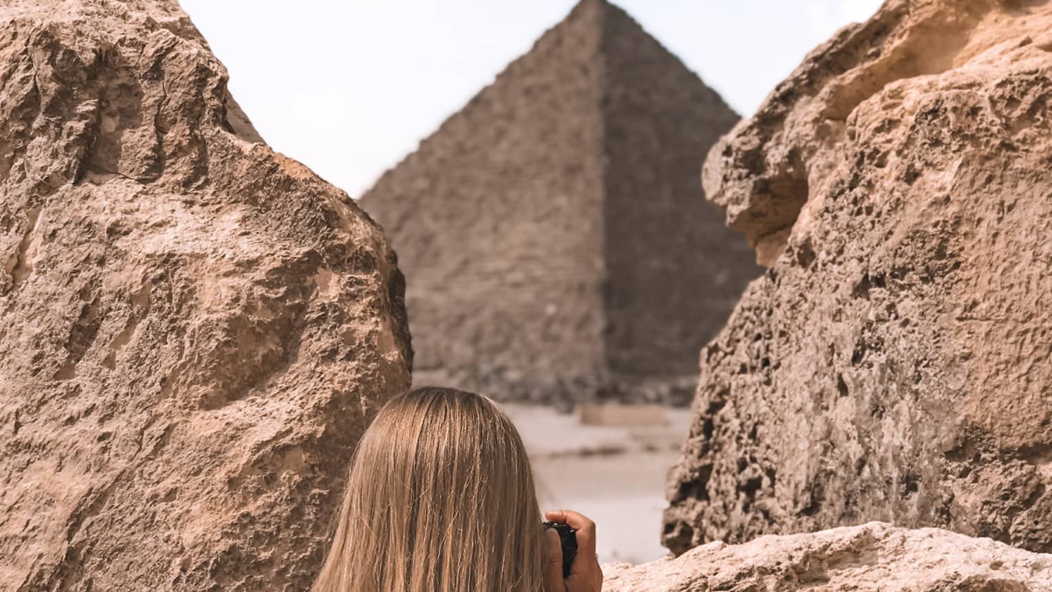 A tourist in a floral dress stands between two large rocks and aims her camera at an enormous pyramid beyond. 