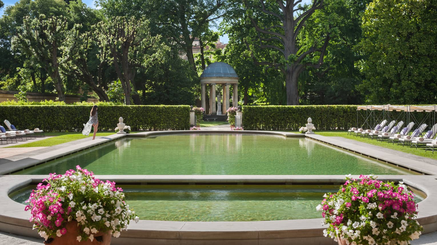 Woman in flowing robe walks past stone ledge of outdoor swimming pool surrounded by gardens