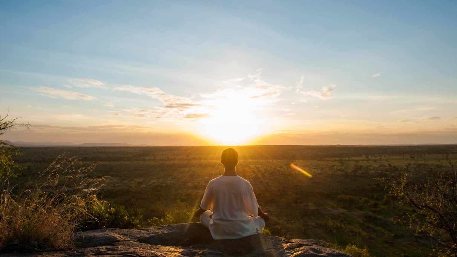 Back of woman sitting, meditating on cliff in front of sunrise 