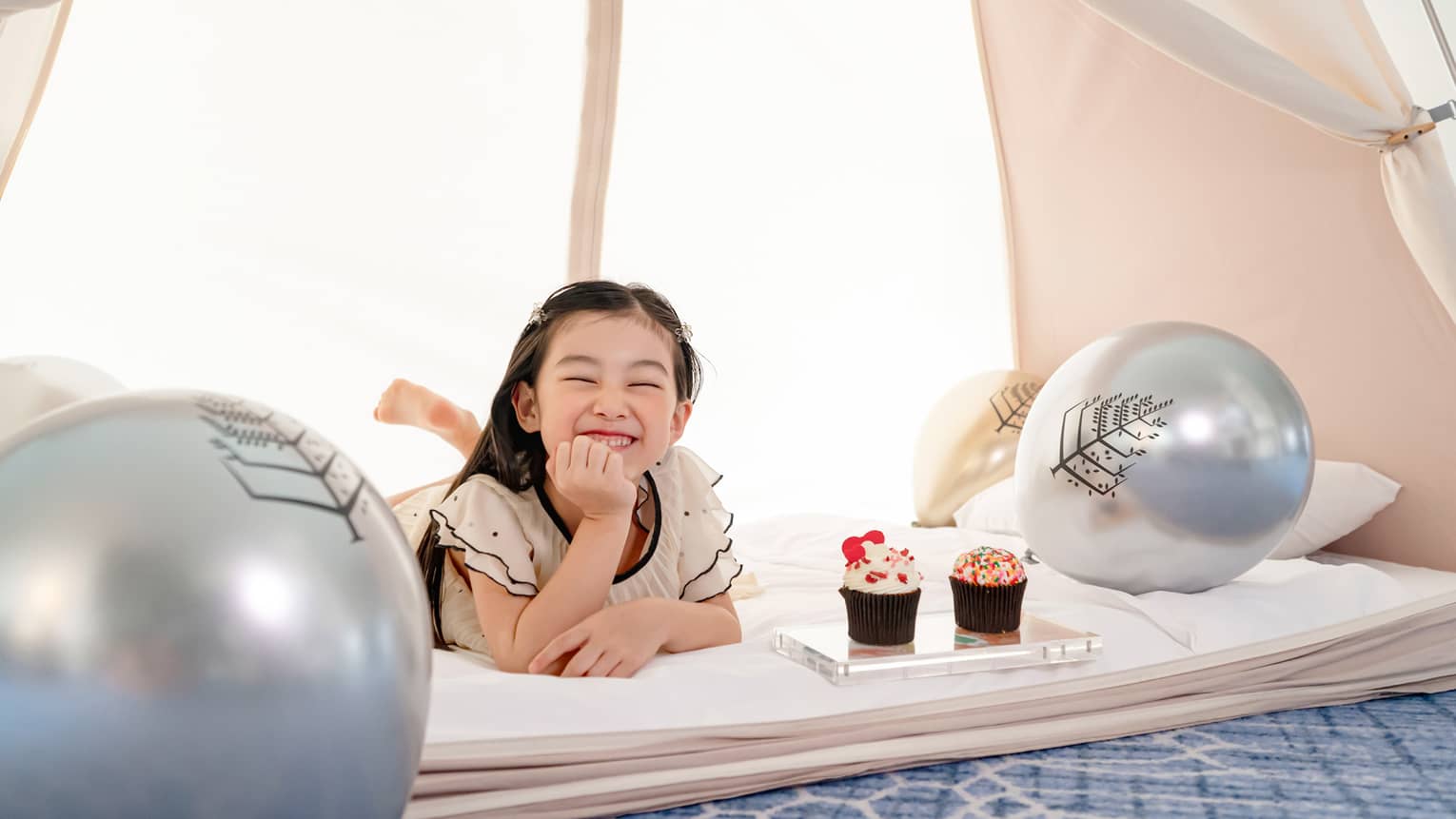 Smiling young girl in in-room kids? tent with gold and silver balloons and cupcakes
