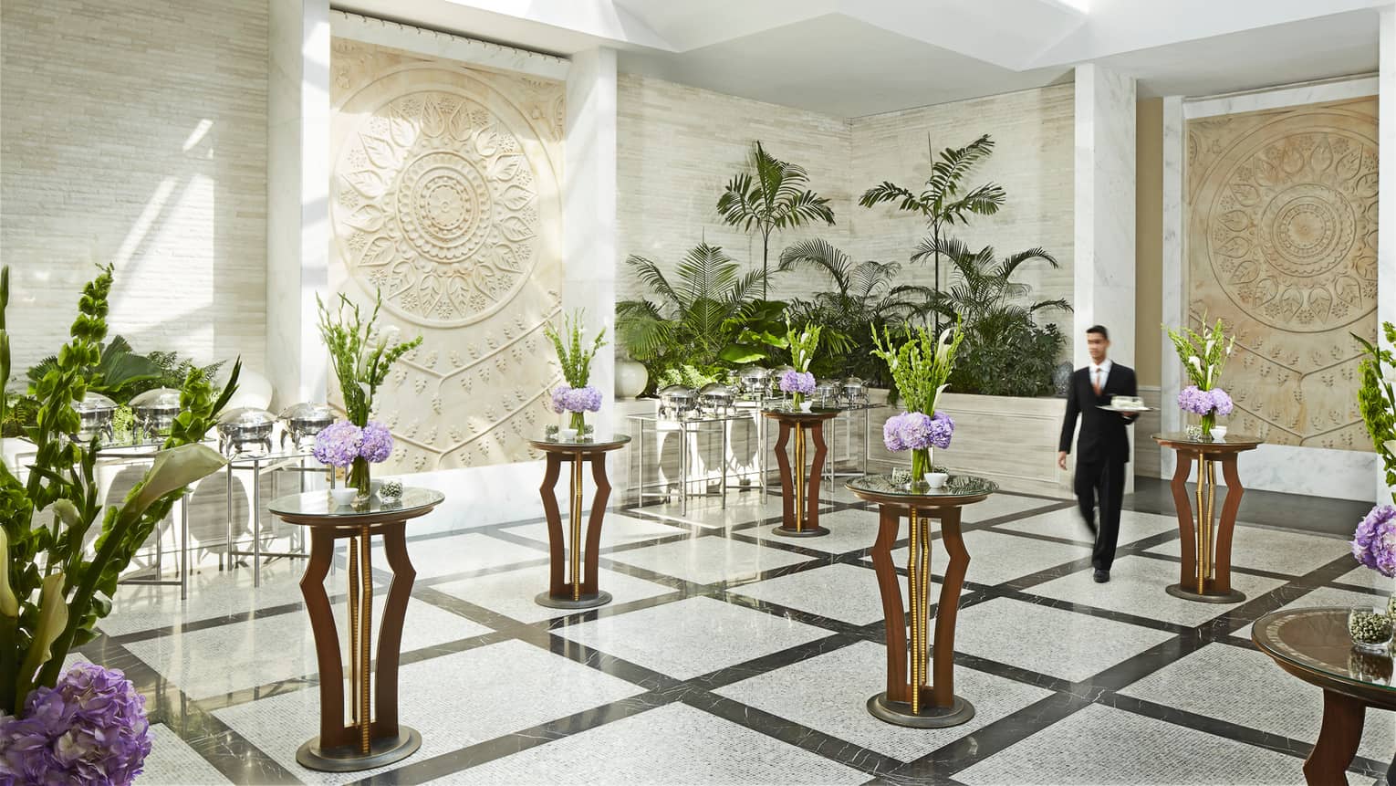 Server with tray walks through white marble room with accent tables, floral displays