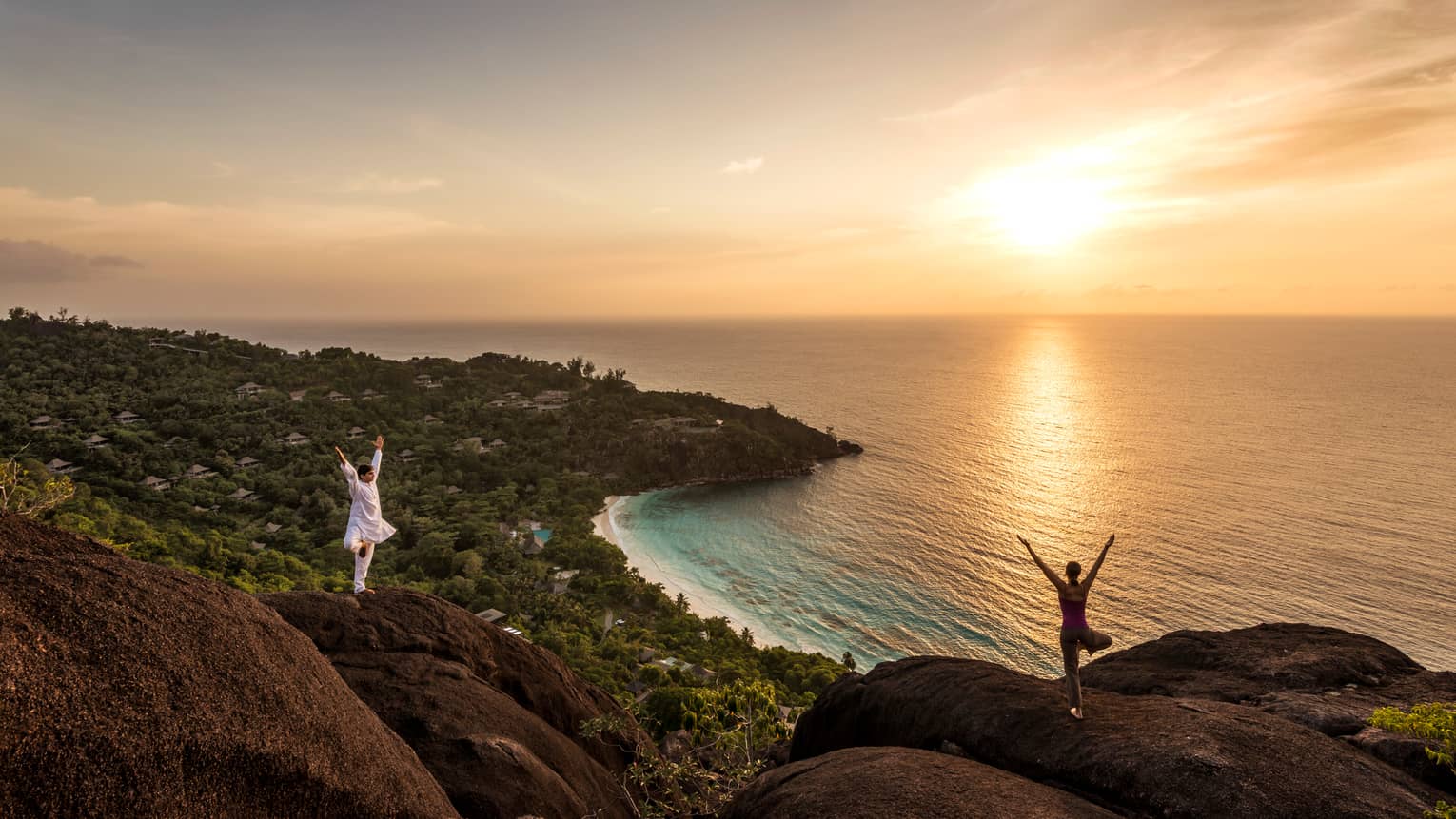 Woman and man stand apart on large boulders overlooking canopy of trees, ocean at sunrise 