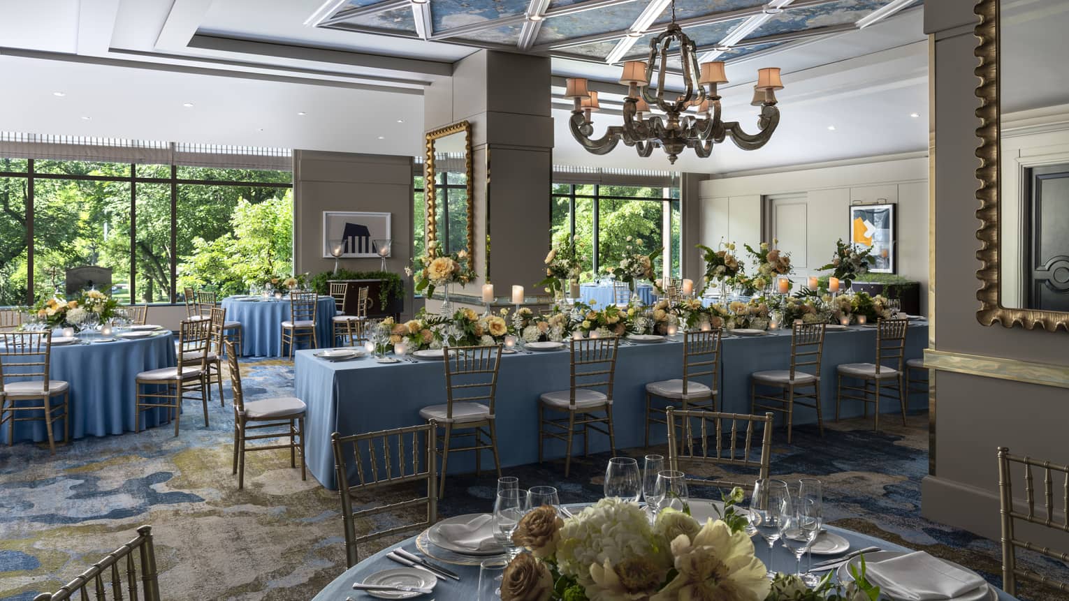 Long and round tables with blue tablecloths with yellow flowers, an old looking chandelier hangs from the ceiling.