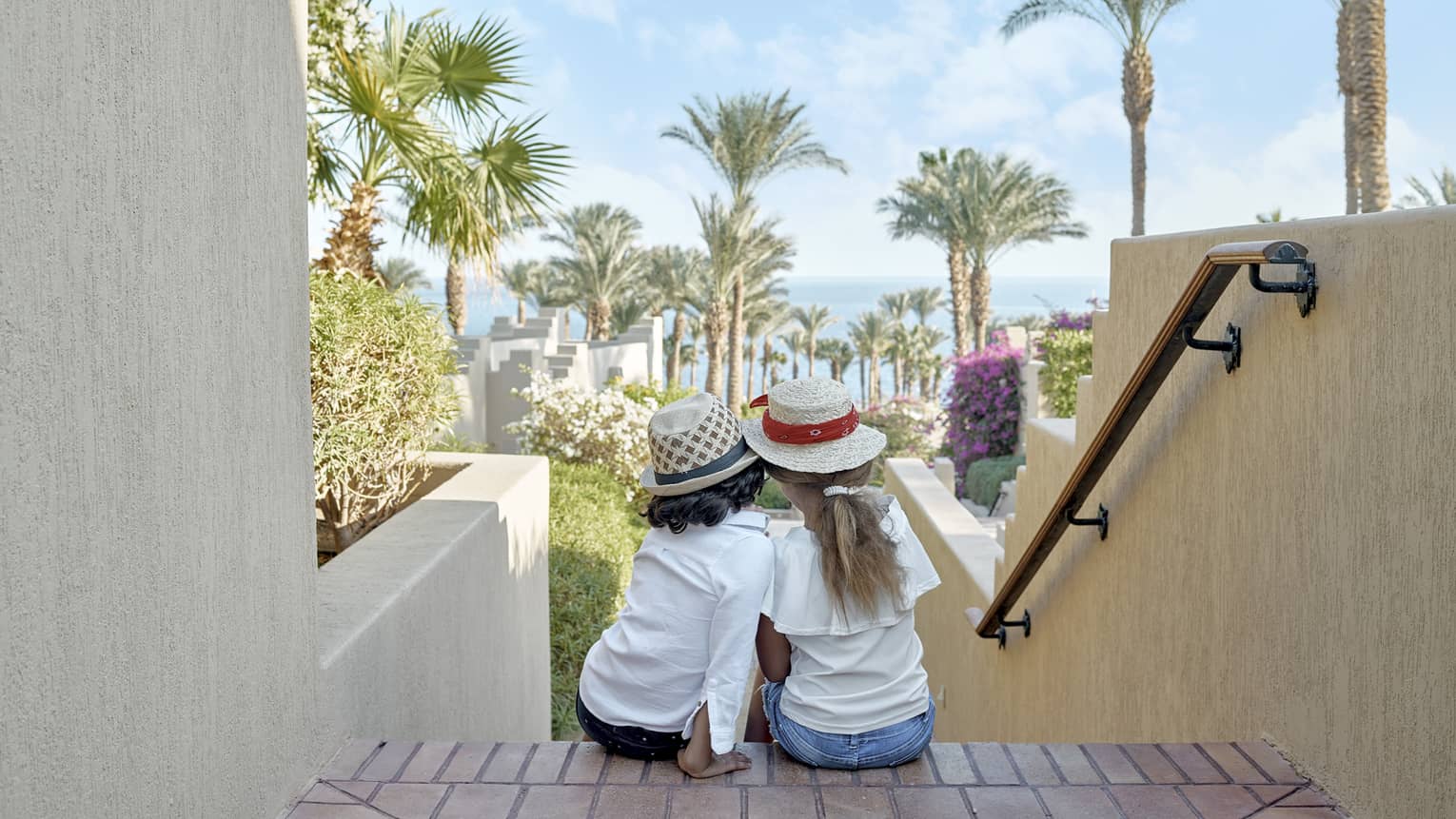 Two young girls sit on stairs looking out to resort grounds with palm trees