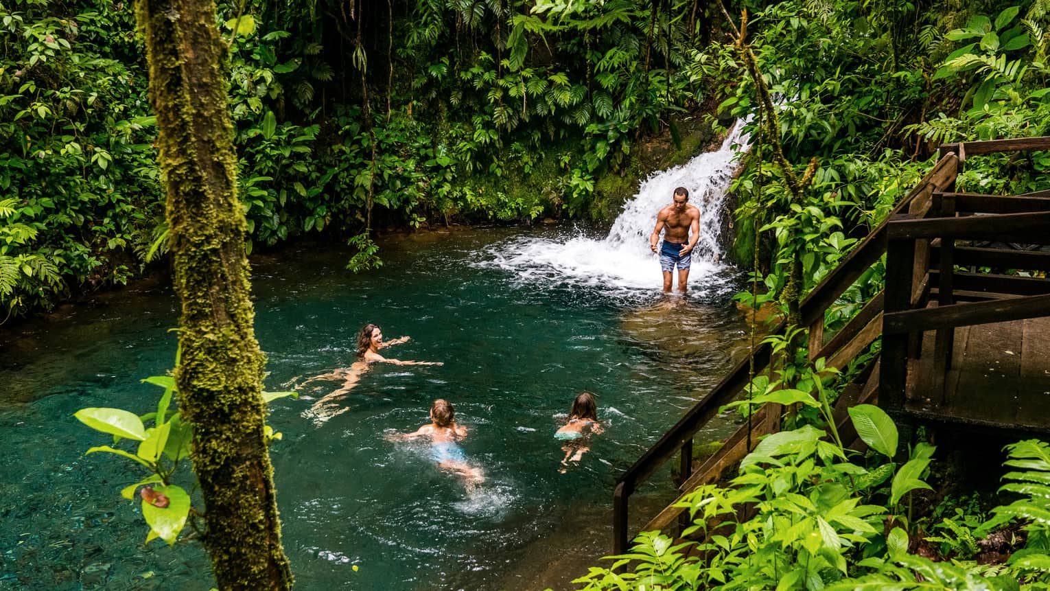 Two adults and two kids frolic at the foot of a waterfall surrounded by lush jungle greenery