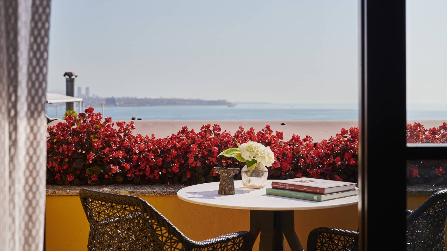 Bistro dining set on orange-walled terrace looking over planter box filled with red flowers to water view