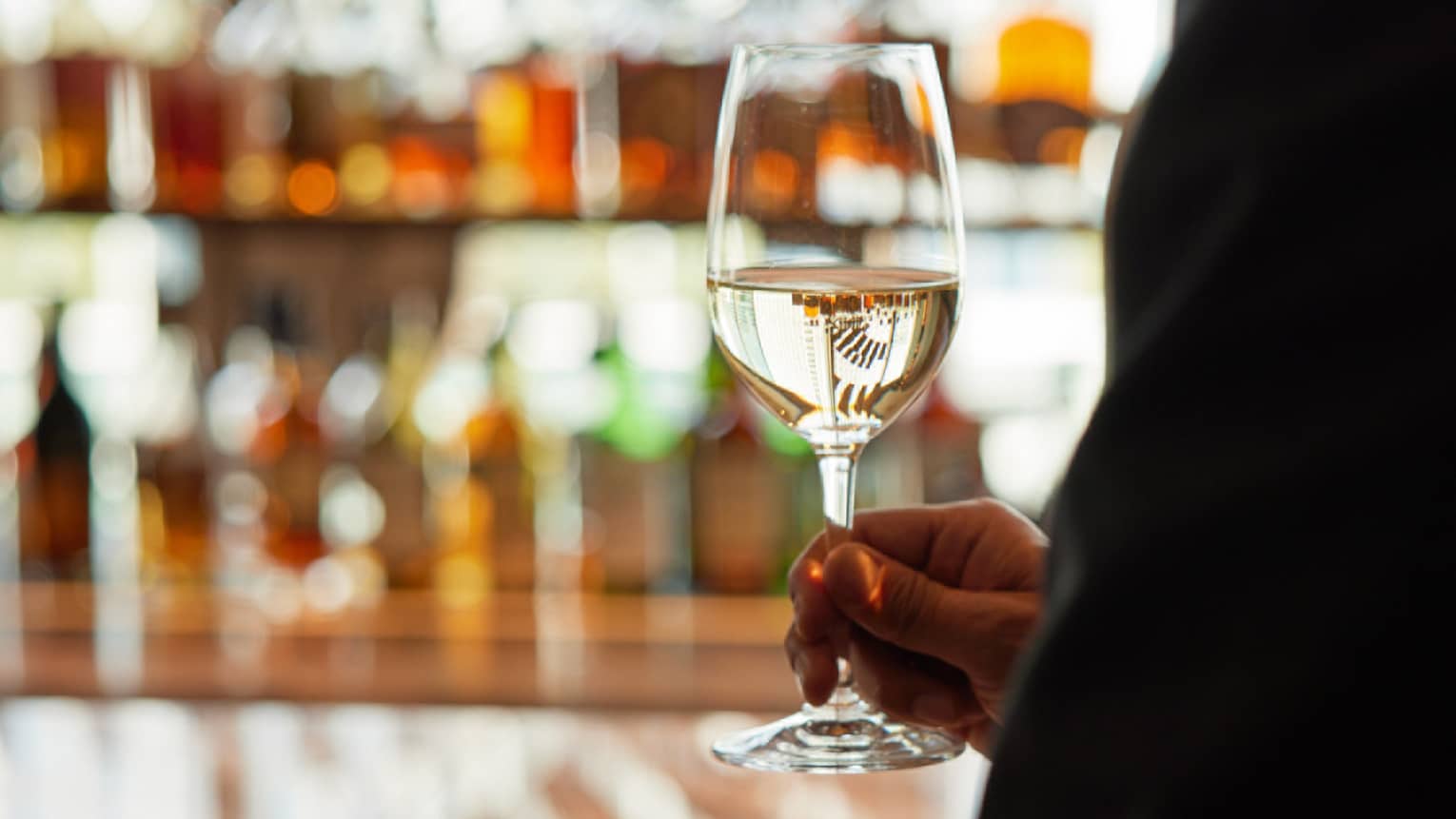 Man holds stemmed glass of white wine against the backdrop of a bottle-lined bar