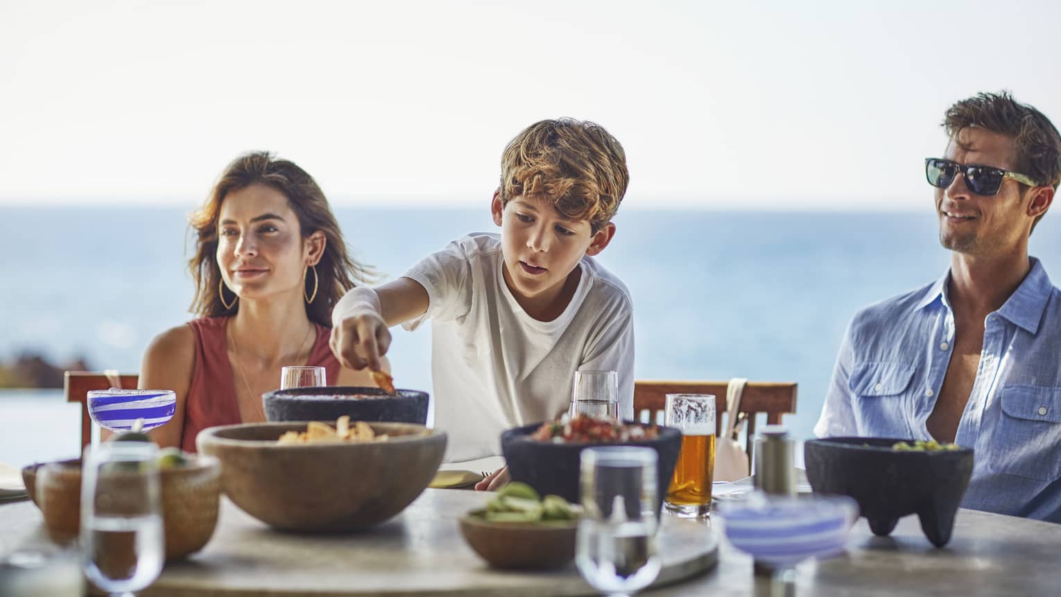 A young boy reaches for a chip bowl on table as a woman and man sit on either side, ocean in the background