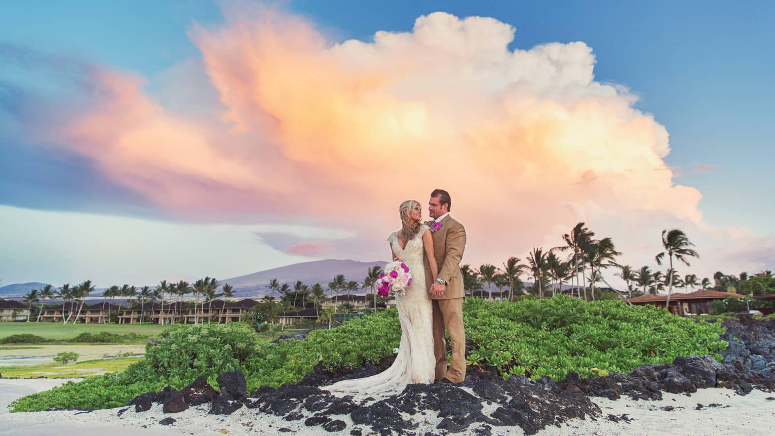 Bride and groom stand on black lava rocks on white sand beach at sunset