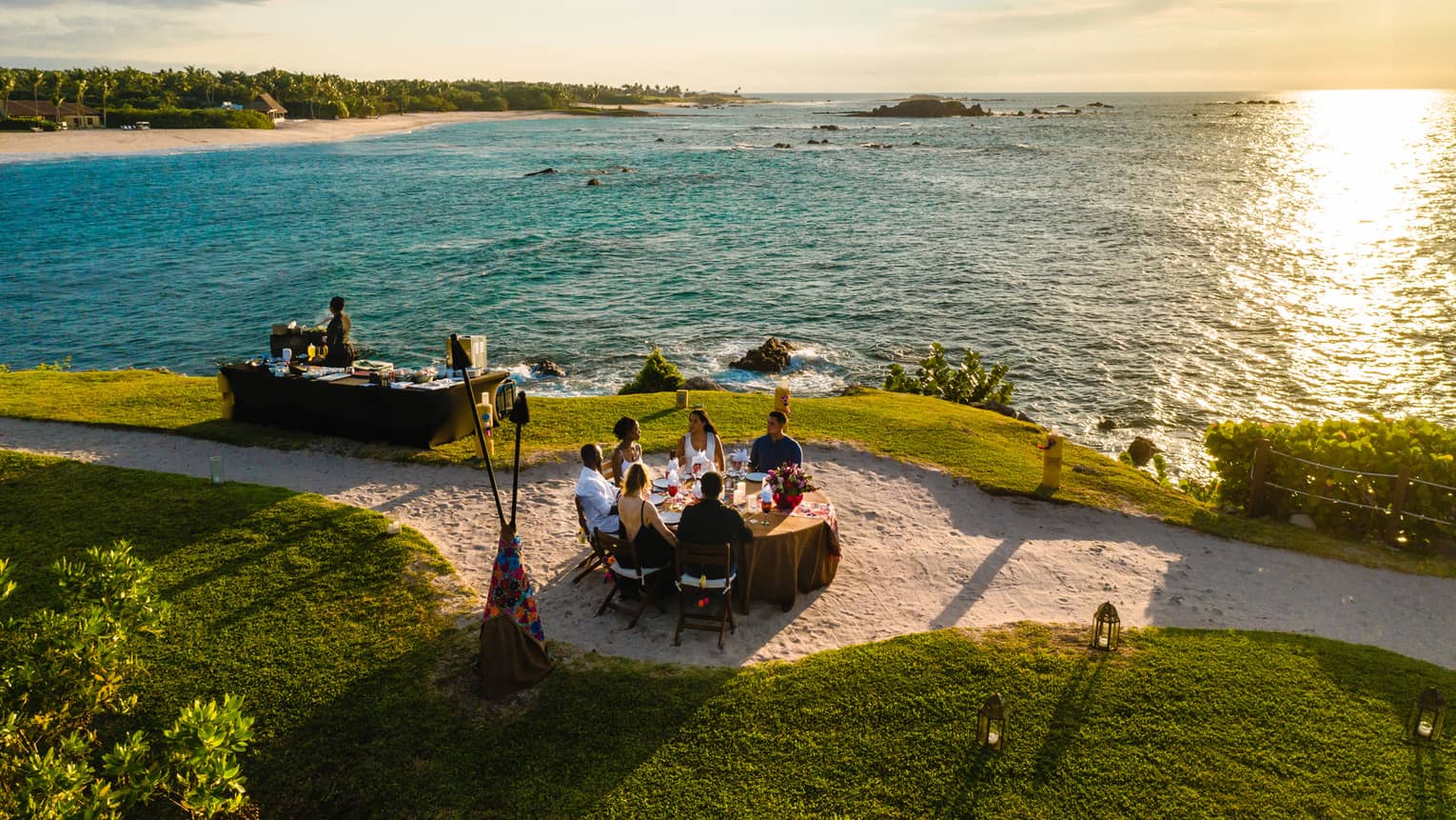 Aerial view of Dinner on the Rock dining table on lawn, sandy path near ocean at sunset