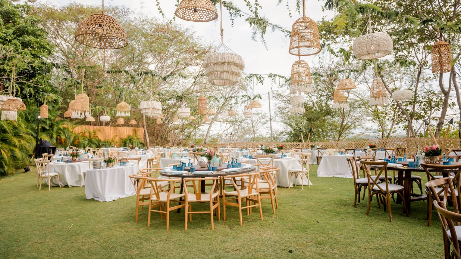 A lawn is set for an outdoor party with various round tables, white table cloths and a variety of straw light fixtures hanging from vined ropes overhead